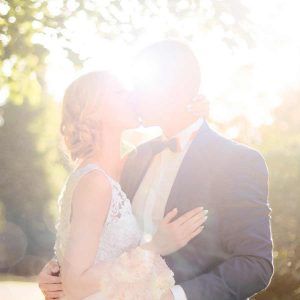 bride and groom kissing near a tree, leaves blurred above and behind them with the bright sun behind them.